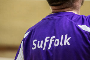 A badminton player stands with their back tot he camera, they are wearing a purple polo shirt with the words Suffolk emblazoned across their back.