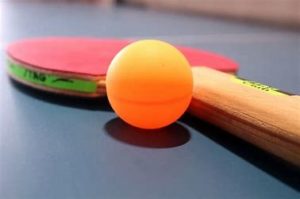 An orange ping pong ball sits next to a red pin pong bat on a blue table tennis table