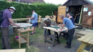 Several mad stand around a shed , they are cutting pieces of wood and making bird boxes.