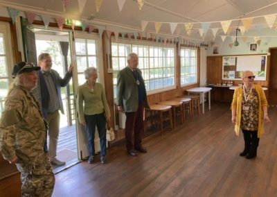 The High Sheriff visits the Cress Pavoilion. He strands in the doorway and admires the room. Bunting can be seen hanging across the ceiling and there is a dark wooden floor. Tables and seats can be seen around the edges of the room