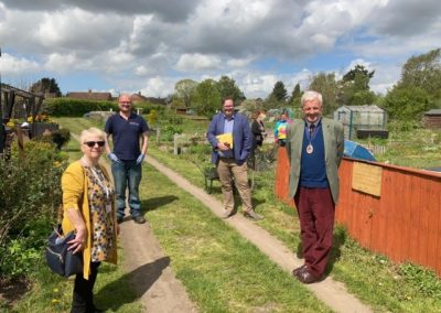 The High sheriff visits the Active Gardens. 4 people stand on a path and smile at the camera. They are surrounded by the plants and vegetable patches of the Active Gardens site.