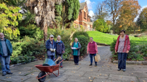 The volunteers of Sudbury Gardens stand proudly in front of a red wheel barrow at the gardens