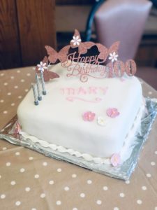 A photo of Mary's 100th birthday cake. Three cancdles stand on top of a white iced cake with a cake decoration that says happy 100th birthday.