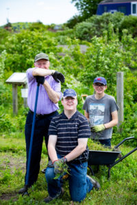 A group of three gardeners from the People's Community Garden smile at the camera as they stand by a wheelbarrow full of garden produce.
