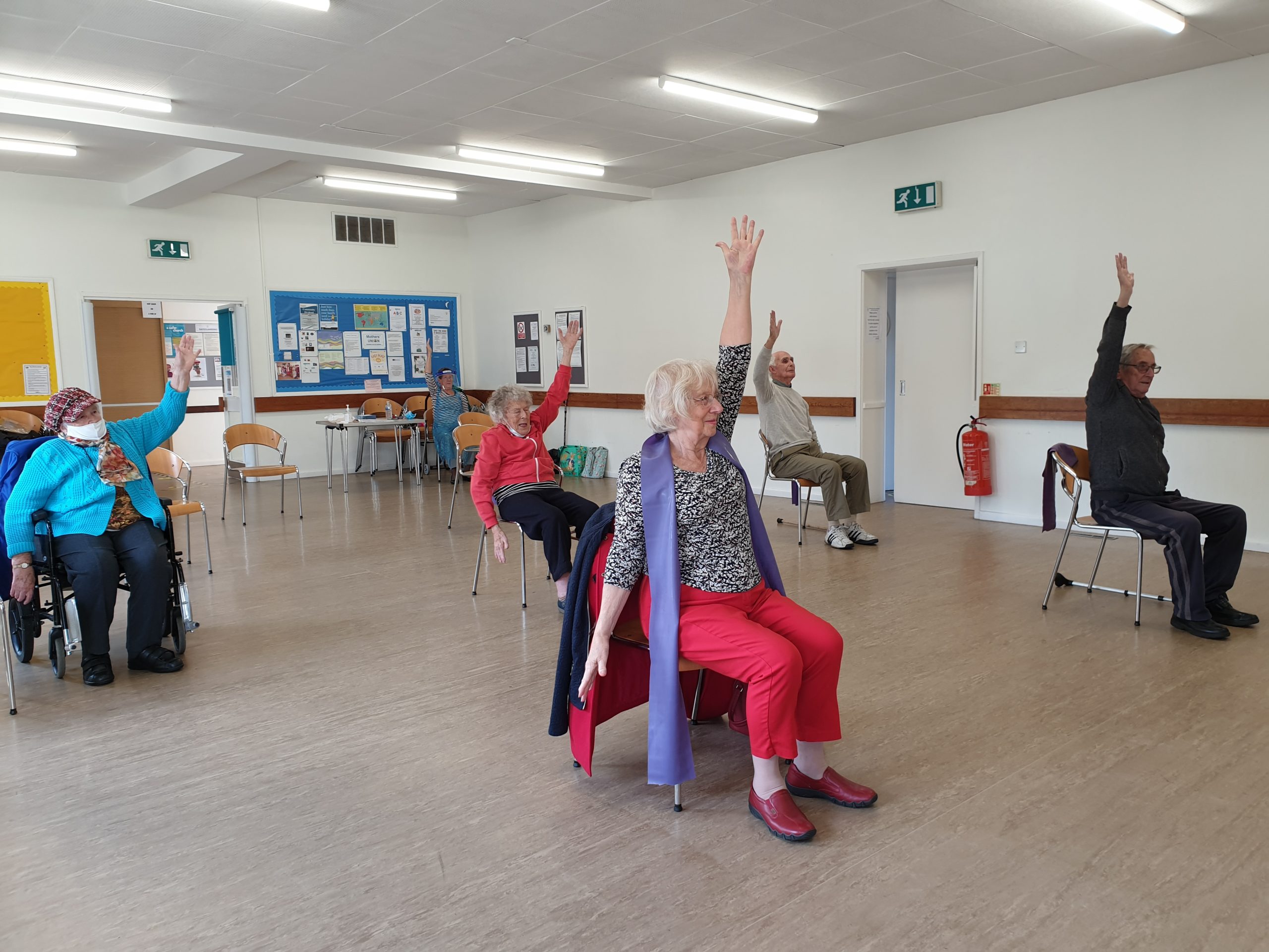 5 people take part in a better balance session, they sit on chairs and raise their arms towards the ceiling.