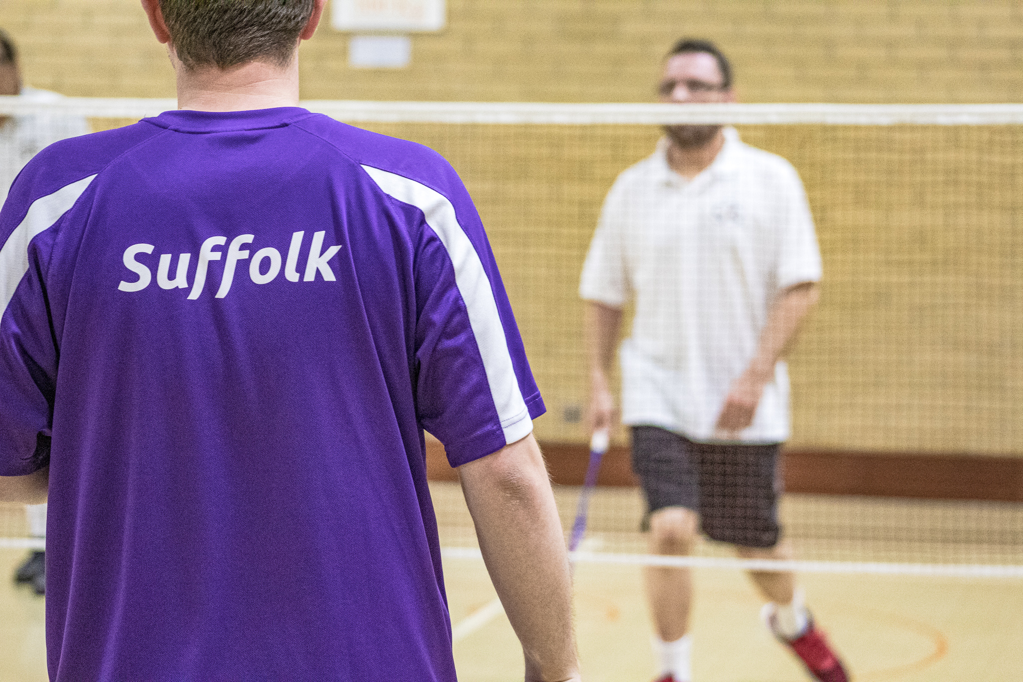 2 people play a game of badminton. One player wears a purple t-shirt with white stripes on the sleeves with the word Suffolk emblazoned on the back of the shirt, he stands with his back to the camera. The badminton net stretches across the middle of the photo and the second player is in the background of the photo. He wears a white t-shirt, black shorts and red trainers. He holds a pruple badminton racquet in his right hand.