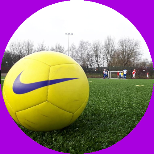 A yellow football sits on a green pitch. A game of walking football can be seen in the background.