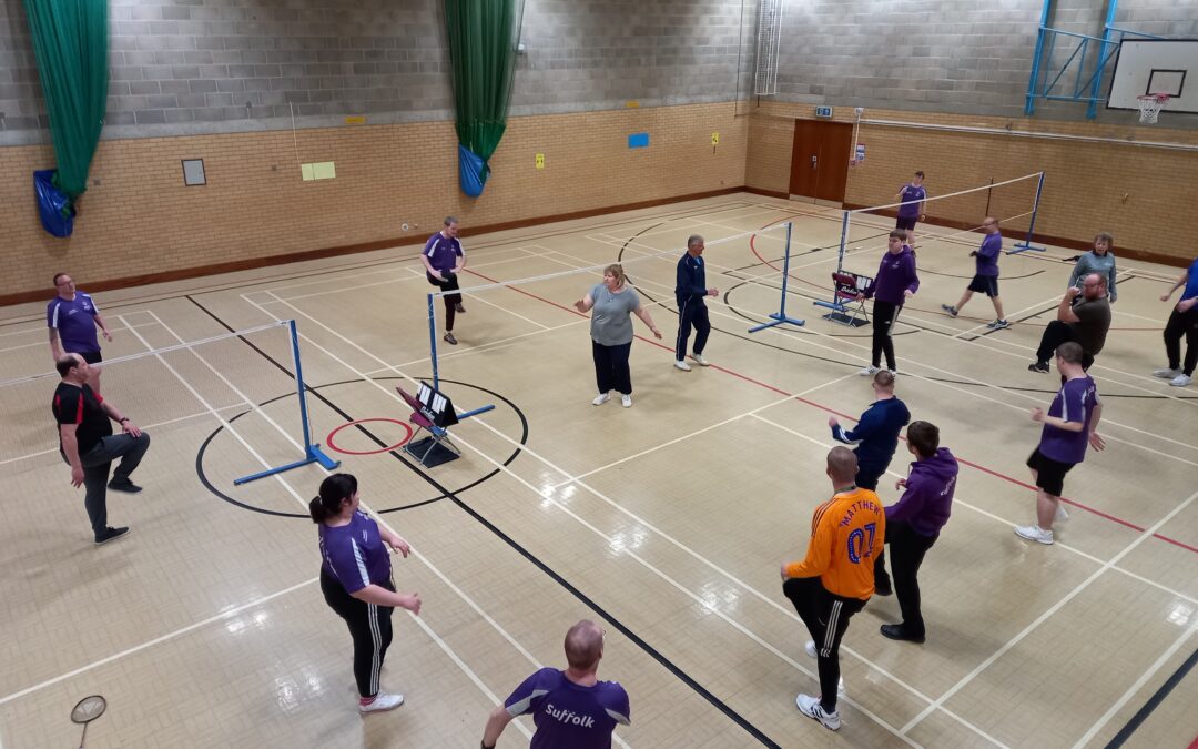 A photo from the badminton tournament. Several players stand on the badminton courts and stretch their legs as they warm up before they play. Everyone wears a purple t-shirt apart from one person who is in an orange top and this person leads the warm up. Several badminton nets can be seen in the photo.