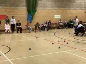 A game of Boccia is in progress. The two teams sit in a row and throw their balls onto the court. The court in front of them is covered in red and clue boccia balls. They all aim for a white jack ball