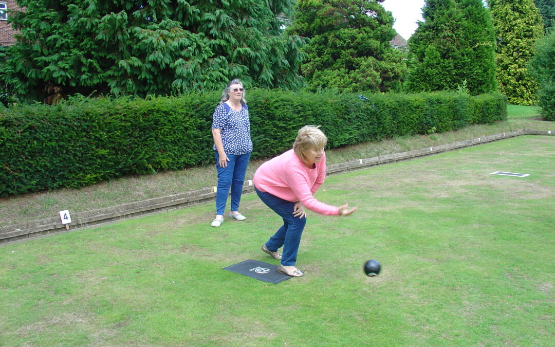A lady in a pink jumper stands at the edge of a bowls green and throws her bowling ball down the green. Another person ion a blue top stands behind her and watches.