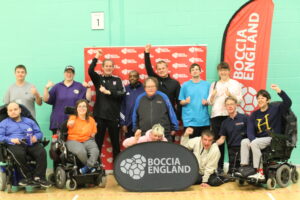 A photo of the boccia team competing in Hatfield. The team all stand and smile at the camera, a few people rasie thier hands in the air in celebration. In front of them is a banner that says Boccia England on it.