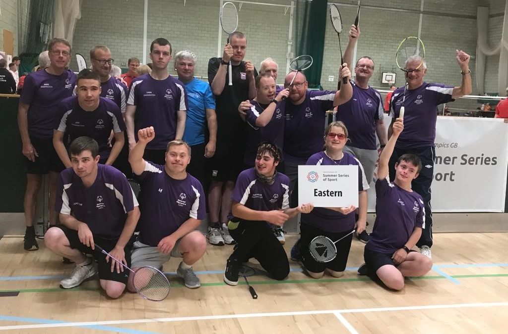 A photo of the team at the inclusive badminton championships in Walsall. The team all wear purple coloured t-shirts and smile at the camera, they all raise their hands in celebration and hold white sign that says Eastern Region on it.