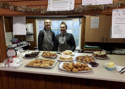 Two people are stood behind a counter, on the counter are different plates of baked goods for sale.