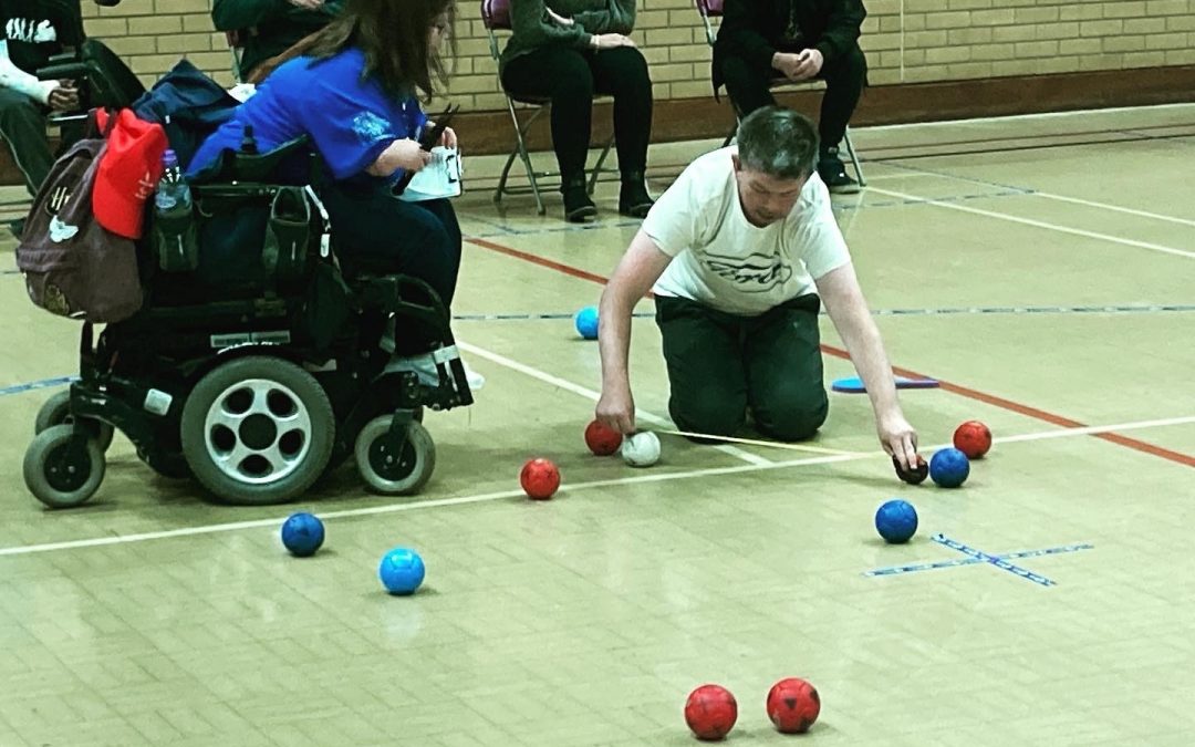 A game of boccia is in play, there is a collection of red and blue balls on the floor and one of the referees takes a tape measure to find out which coloured ball is closest to the white jack ball.