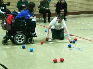 A game of boccia is in play, there is a collection of red and blue balls on the floor and one of the referees takes a tape measure to find out which coloured ball is closest to the white jack ball.