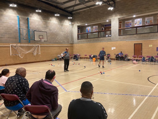 4 people sit in chairs and watch as a game of boccia plays out in front of them, there are red and blue balls sitting on the court as a game is in progress. 2 referees can be seen on the court as the game continues.