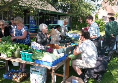 People sit out side the front of hte pavilion, there are table set up selling plants and severl people stand looking at the produce on sale. Other sit at a picnic table and drink tea.