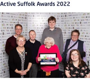 A group of people all stand together and have a photo taken, they all smile at the camera and hold up their runners up certificates that they received at the active Suffolk awards ceremony.