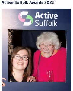 2 women pose in between a blue cardboard frame that says Active Suffolk on it, they both smile at the camera.