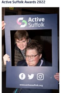 2 men pose in between a blue cardboard frame that says Active Suffolk on it, they both smile at the camera.