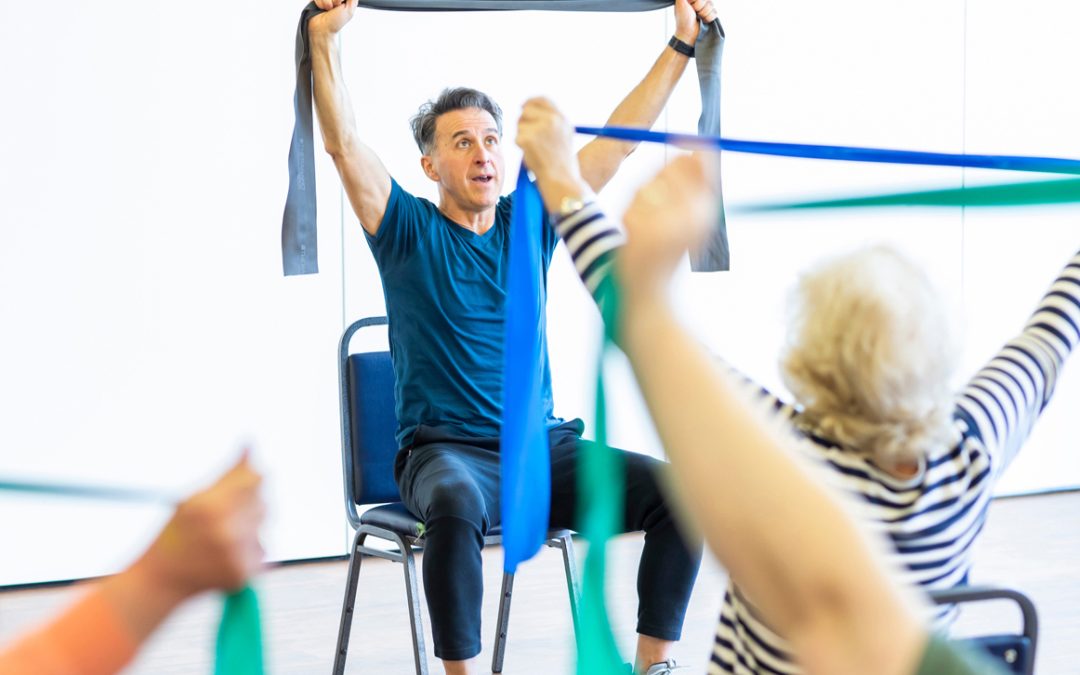 A man holds an elastic exercise band above his head as he does a stretch. He sits opposite a class full of participants who copy his actions.