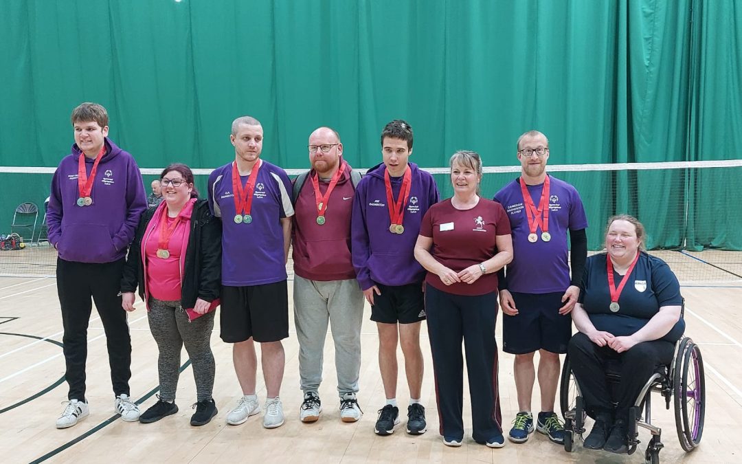 8 people all stand in a row on a badminton court, they are posing for the photo after competing in a badminton tournament. They all wear a competitor’s medal with a red ribbon and gold medallion.
