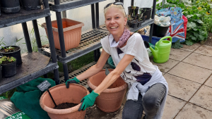 A lady wearing a grey t-shirt and green gardening gloves smiles at the camera. She holds a red flower pot that is half filled with compost.
