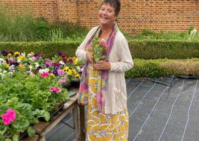 A lady in a yellow dress and white cardigan stands by a table full of plants. She holds a pink flower and smiles at the camera.