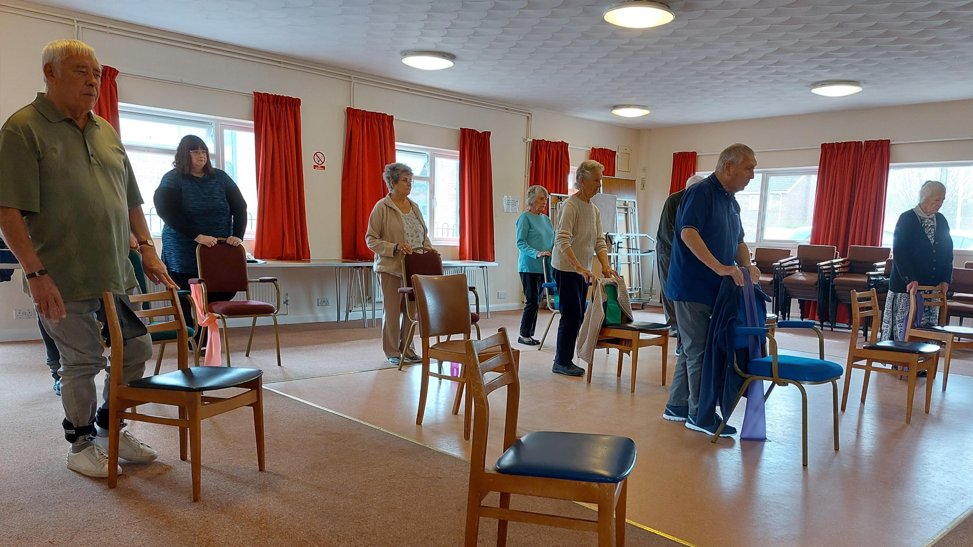 Several people stand in a room, they all lean on chairs as they take part in a Better Balance session