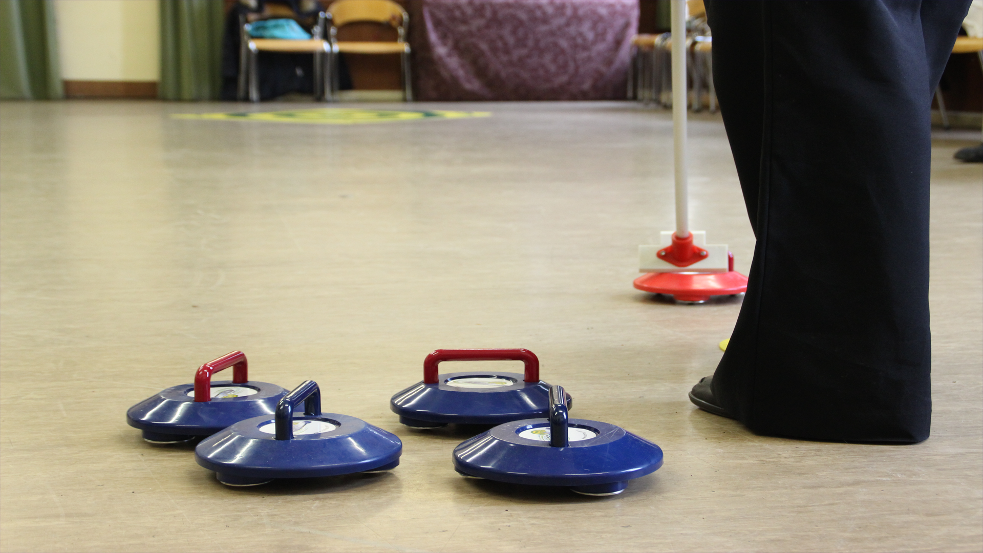 4 blue Kurling irons can be seen on the ground, a person wearing a black pair of trouser and a brush.
