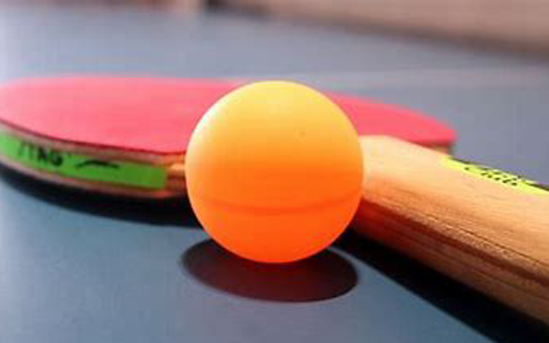 A red ping pong bat lies on a ping pong table. Next to it is an orange ping pong ball.