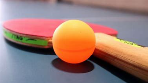 A red ping pong bat lies on a ping pong table. Next to it is an orange ping pong ball.
