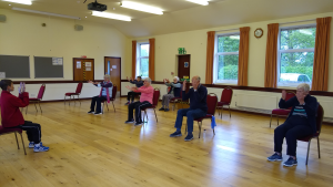 Several people sit on chairs in a village hall. They all raise their arms upwards, stretching and exercising as part of a better balance session.