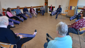 Several people sit on chairs in a village hall. They all have an exercise band around their legs, they each raise their leg in an exercise as part of a better balance session.