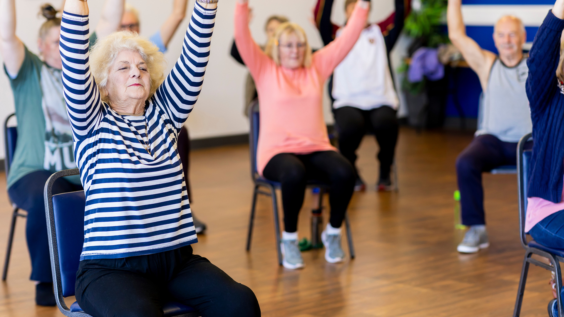 Several people sit on chairs in a village hall. They all raise their arms upwards,, stretching and exercising as part of a better balance session.