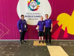 3 people stand in front of a giant curtain that has the special olympics logo on it. They all wear blue polo shirts and smile at the camera.