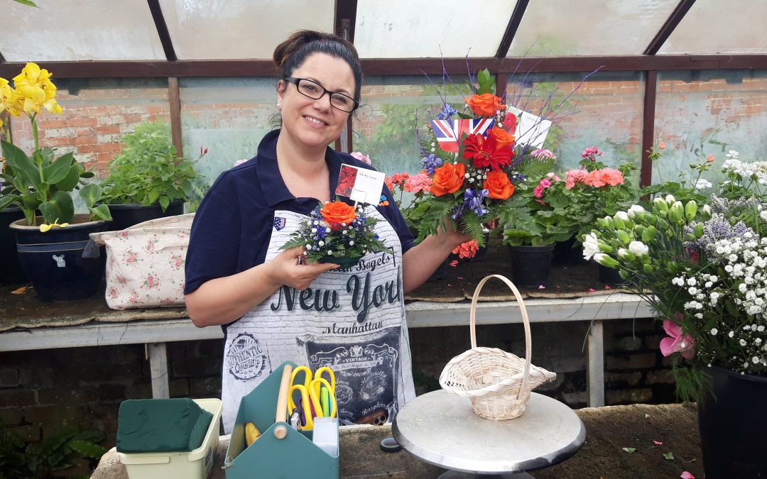 A lady with brown hair and a blue t-shirt smiles at the camera and holds up a flower arrangement. On the table in front of her is a display basket and some equipment she uses to create her flowerdisplays.