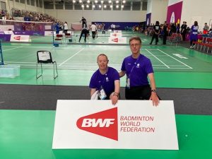2 men in blue t-shirts stand behind a world badminton sign.