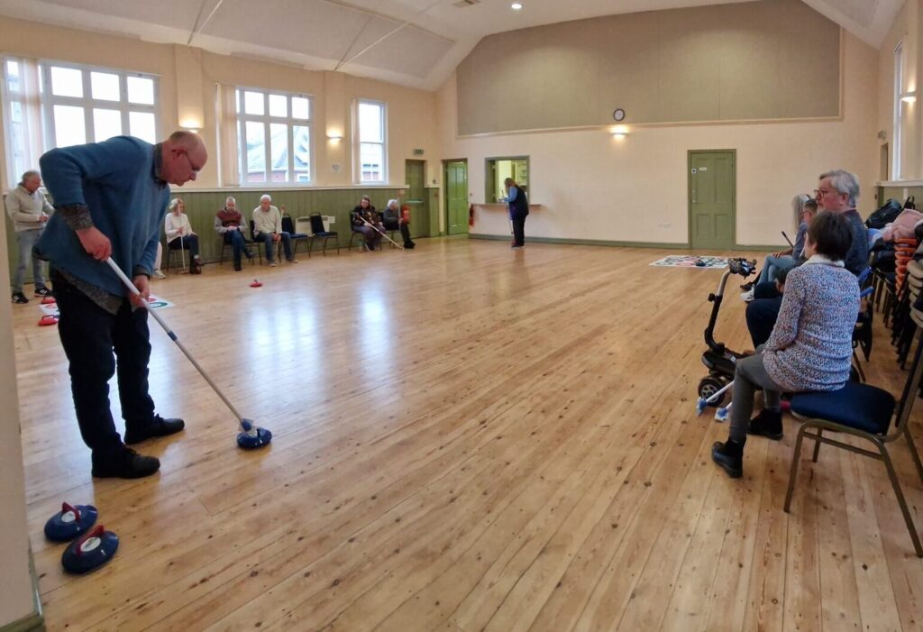 A man playing kurling with members as audience in a hall