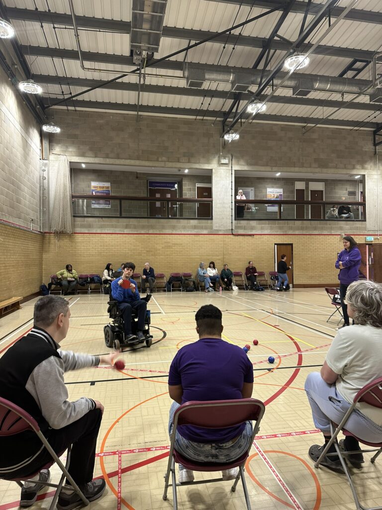 A man participant throwing a boccia ball with athletes, volunteers and audience watching
