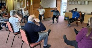 A group of participants with the instructor doing a chair based exercise inside a hall